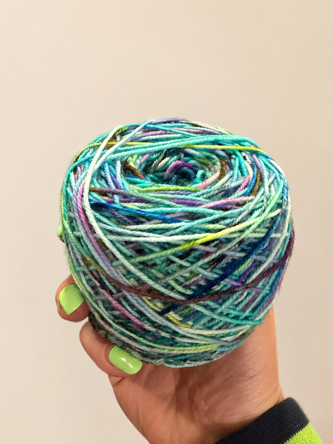 A hand with lime green nail polish holds a caked ball of variegated hand-dyed yarn with shades of teal, blue, green, purple, and yellow against a plain beige background.