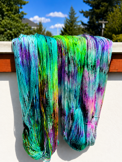 Two large hanks of colorful hand-dyed yarn hanging outdoors against a white wall, with trees and a blue sky visible in the background.