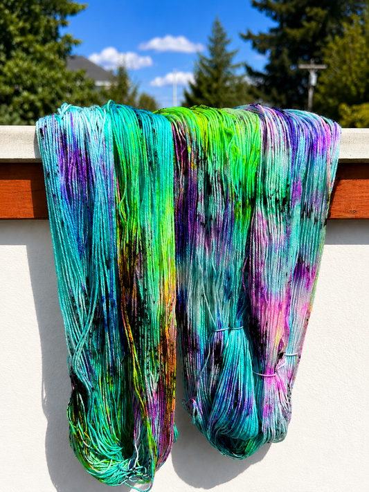 Two large hanks of colorful hand-dyed yarn hanging outdoors against a white wall, with trees and a blue sky visible in the background.