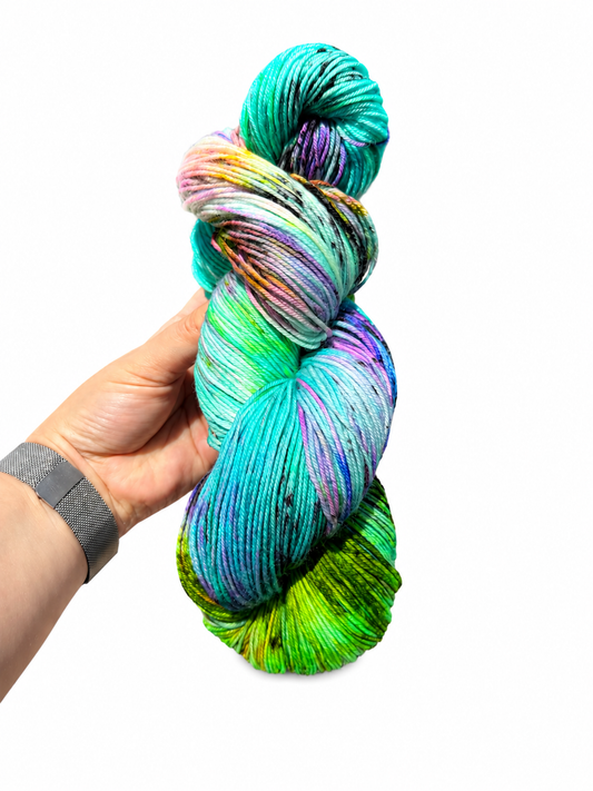 A hand holding a twisted skein of hand-dyed variegated yarn with vibrant shades of teal, green, purple, pink, and yellow on a plain white background.