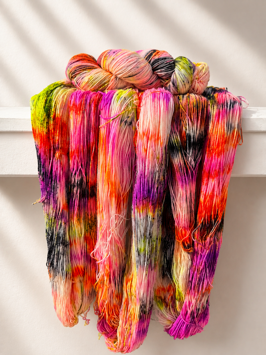 A bundle of colorful skeins of hand-dyed yarn draped over a white shelf, featuring vibrant shades of pink, orange, purple, green, yellow, and black in a tie-dye pattern.