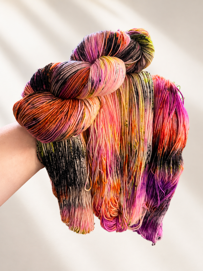 A hand holding a bundle of four skeins of variegated hand-dyed yarn with a rainbow of colors including pink, orange, purple, yellow, and black, set against a warm off-white background with soft leaf shadows.