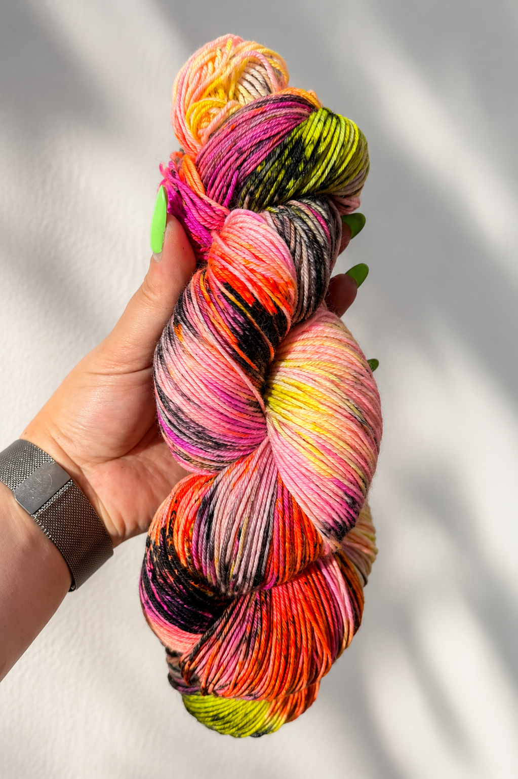 A hand holding a twisted skein of variegated hand-dyed yarn with bright pink, orange, yellow, green, and black flecks, set against a white wall with soft leaf shadows.