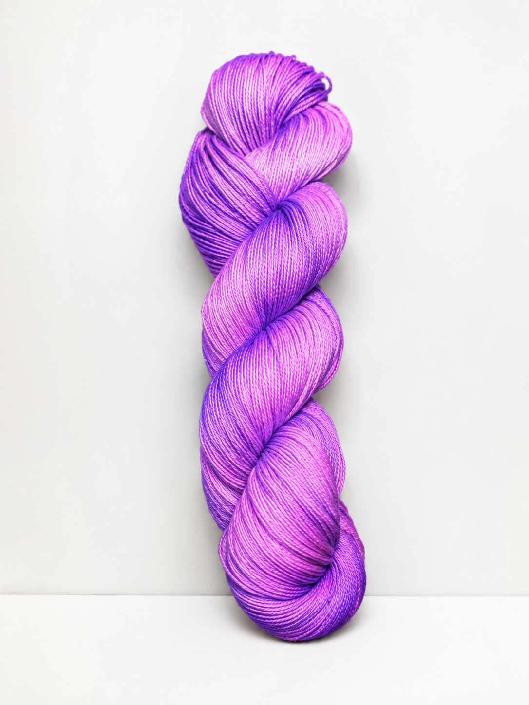 A single skein of twisted hand-dyed yarn in a vibrant purple and magenta gradient, standing vertically against a clean white background.