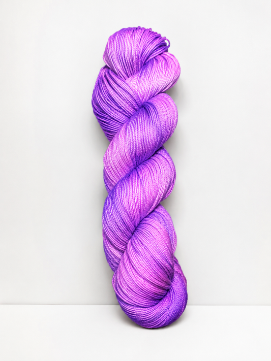 A single skein of twisted hand-dyed yarn in a vibrant purple and magenta gradient, standing vertically against a clean white background.